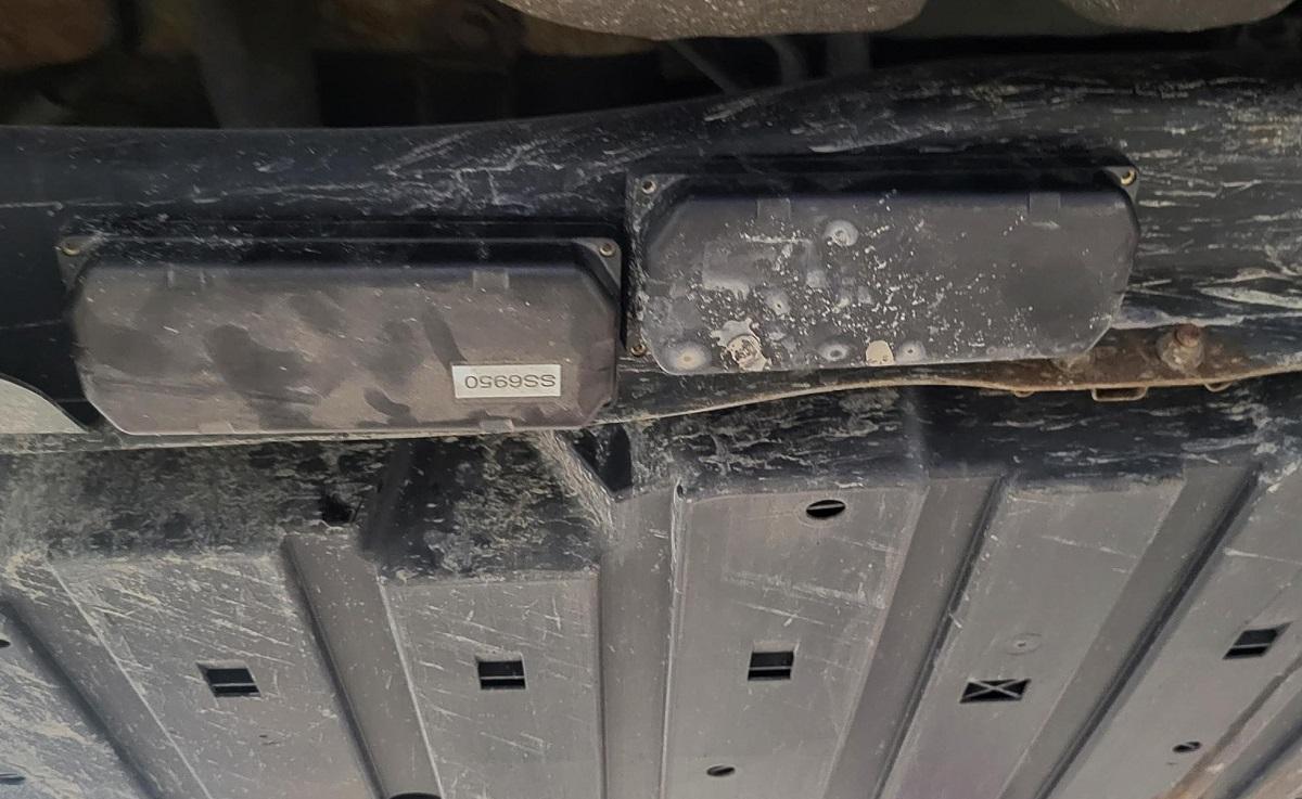 Two GPS trackers, rectangular black plastic boxes similar in size to a deck of playing cards, magnetically attached to the underside of a car.