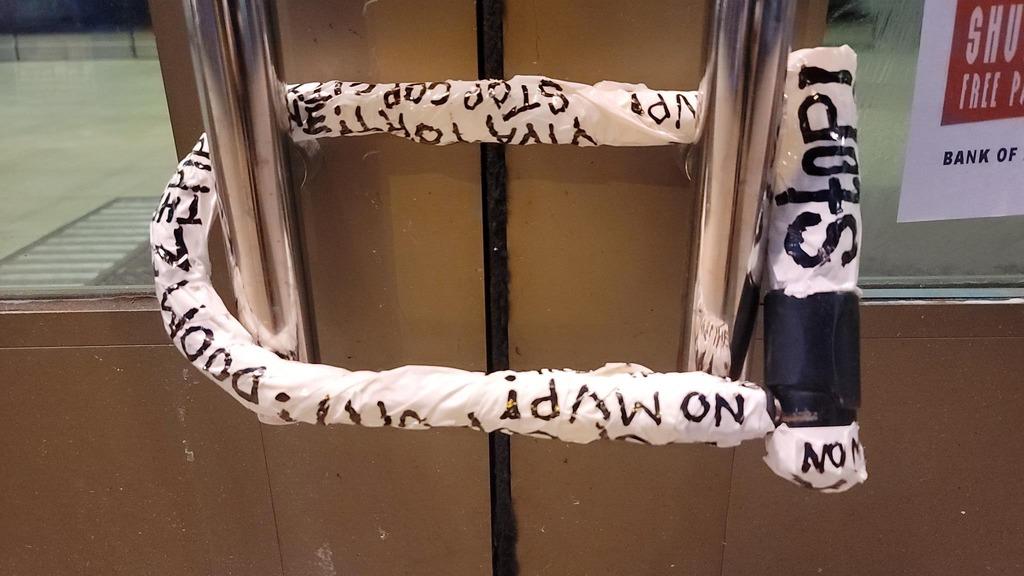 A bicycle u-lock is locked to the front doors of a Bank of America branch. It is covered in white tape with various slogans written on it: No MVP! Stop Cop city! Viva Tort! Doom 2 the Pipeline! Stop!