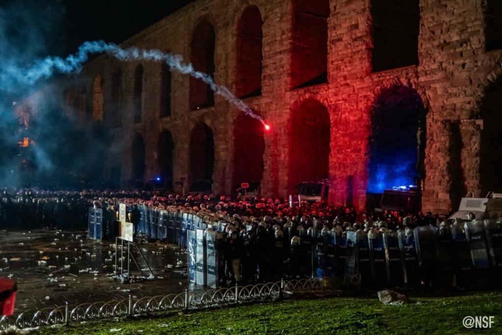 A dense crowd of hundreds of riot police with shields stand in front of multiple stone archways at night. A single red flare arcs over the police, thrown by someone out of frame.