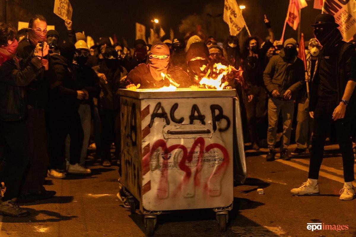 A masked protestor pushes a small burning dumpster with "ACAB" and "1312" spraypainted on it. They are surrounded by other masked protestors.