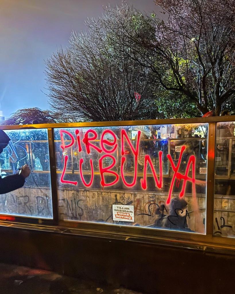 A variety of graffiti is written on walls and transparent dividers, most visibly red text which translates to english as “Queer Resist”