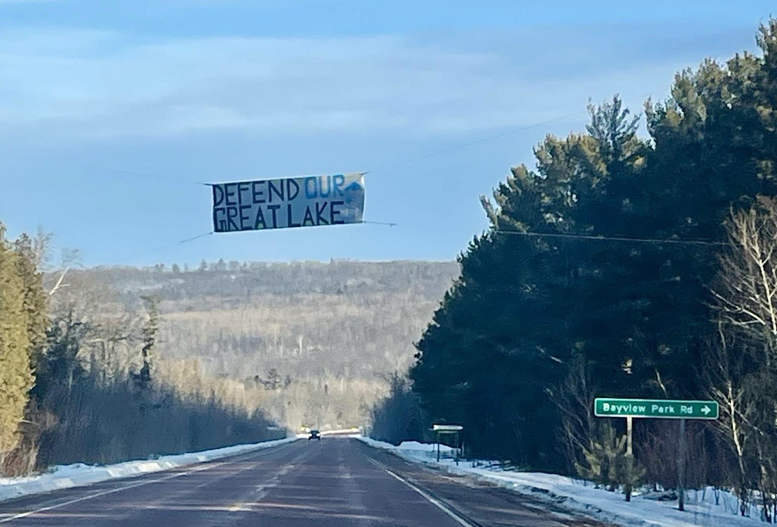 A banner reading "Defend Our Great Lake" hangs suspended over an interstate. The ground is snowy and the area is heavily forested with pine trees.
